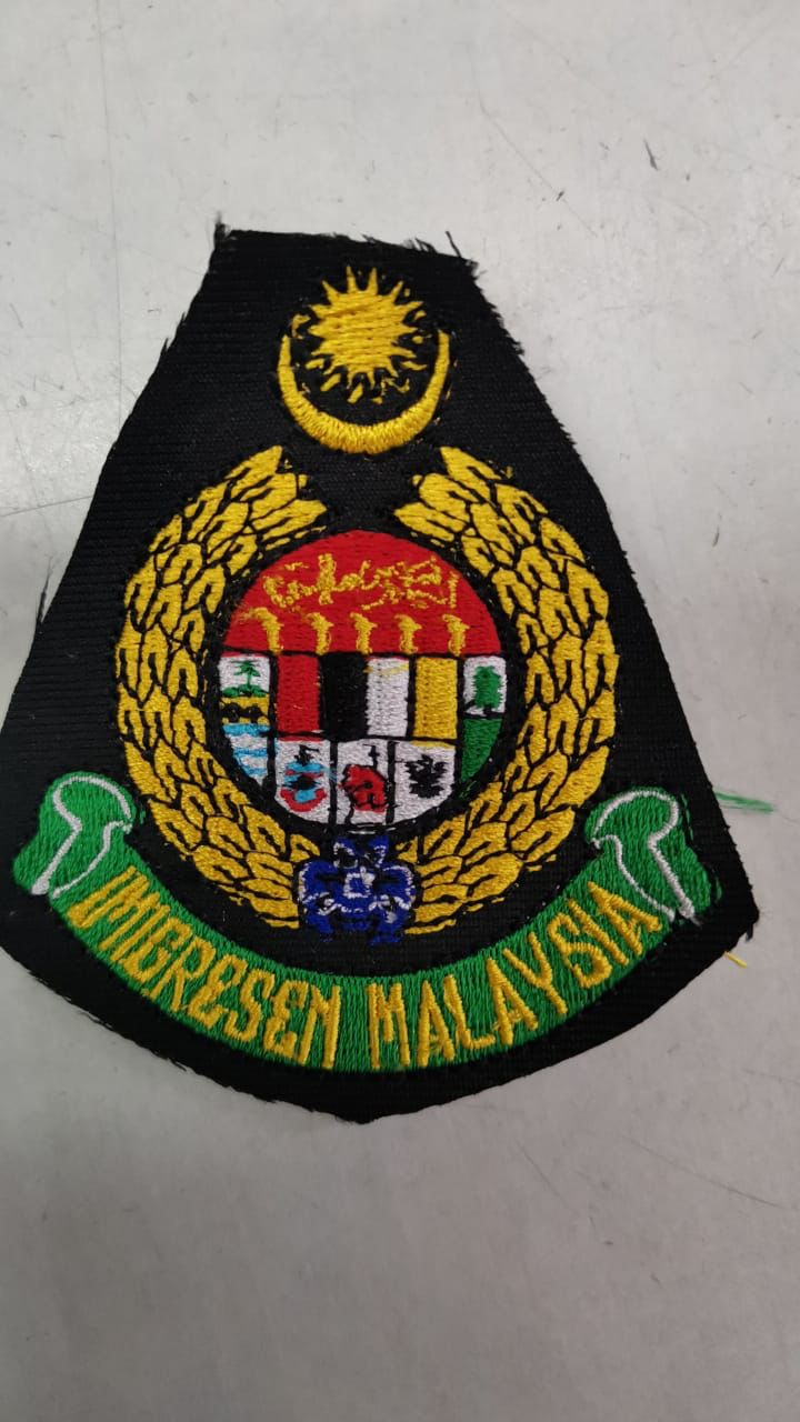 Lencana bersulam Imigresen Malaysia pada kain hitam di atas meja putih